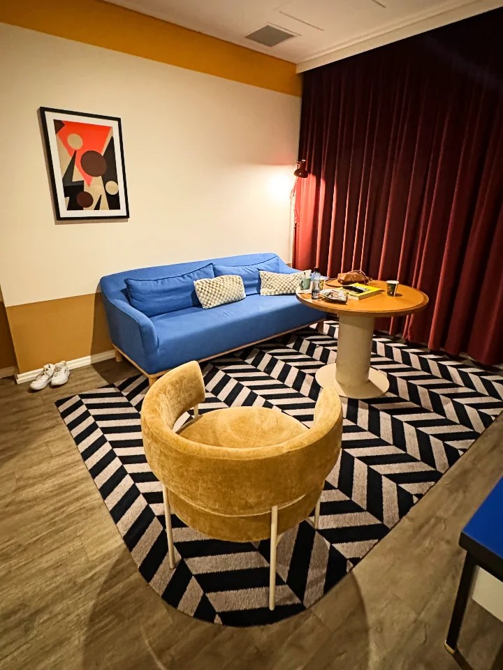 A cozy hotel room with a blue sofa, a round table, and a patterned rug. The walls feature modern art and curtains are drawn in a rich burgundy color.