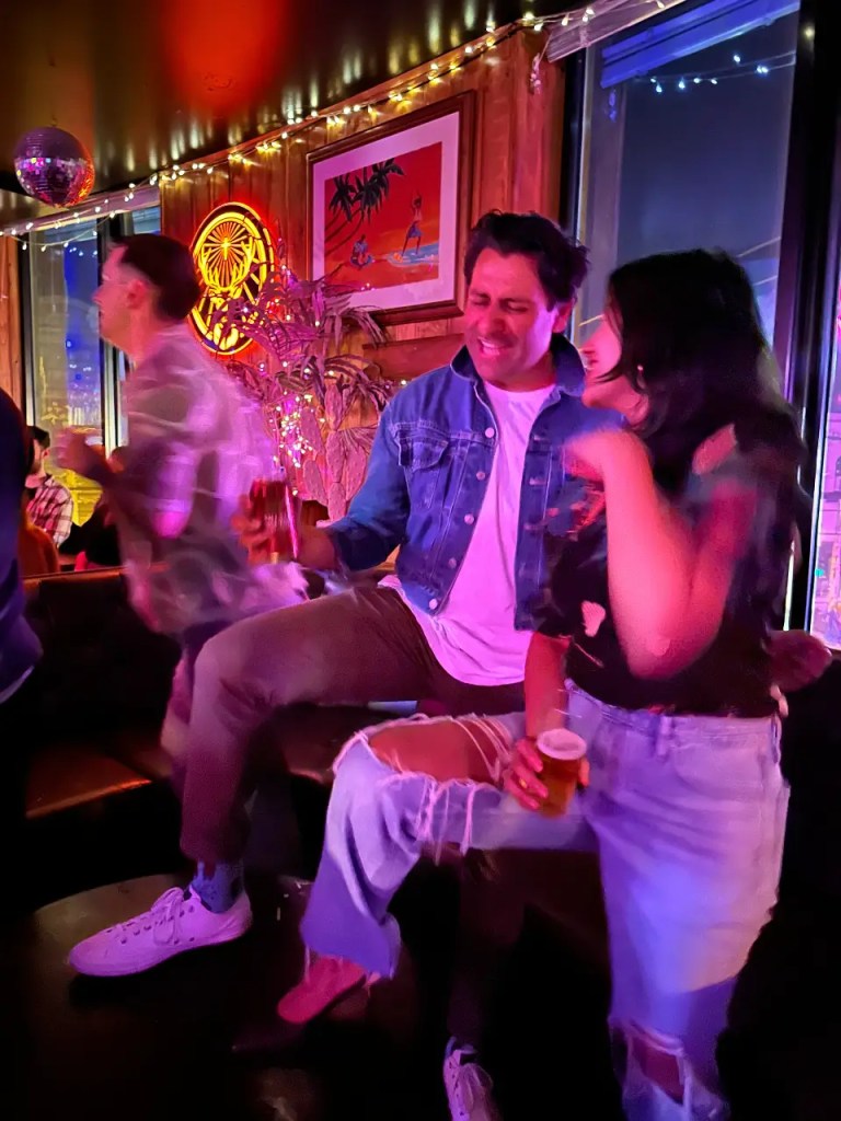 A scene of two friends enjoying drinks in a bar, surrounded by colorful lights and decorations. One has a playful expression, while the other is standing close by, laughing. Both appear to be having a great time.
