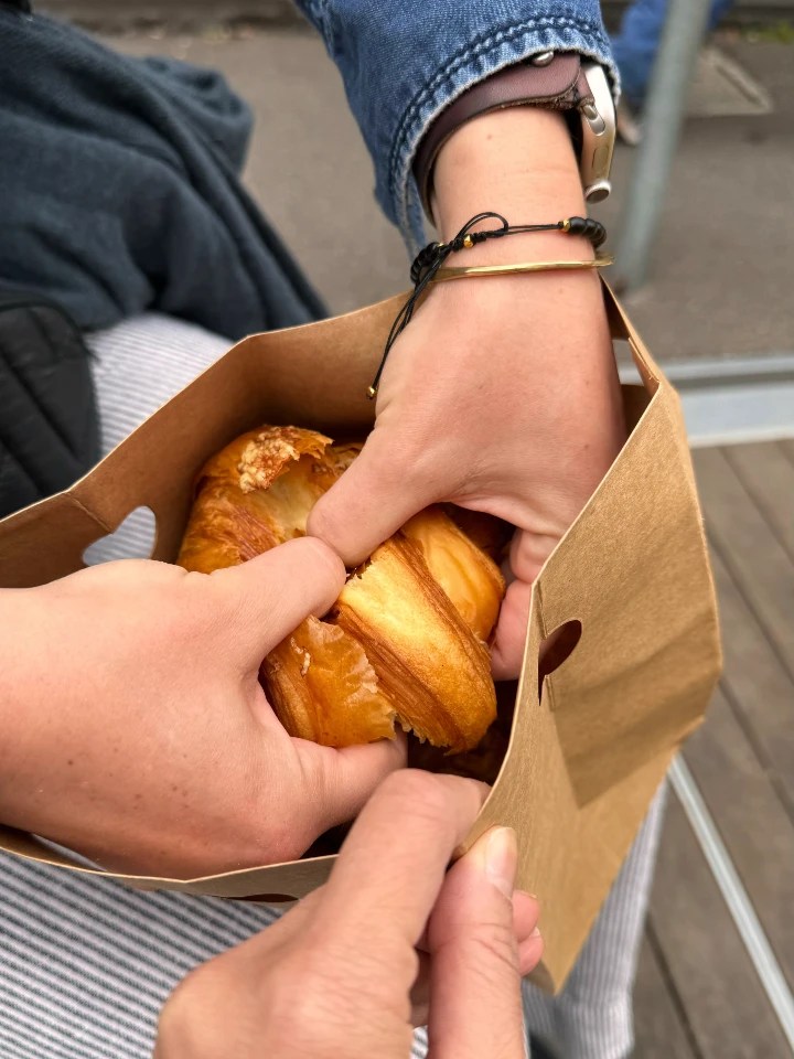 Two hands reaching into a paper bag to grab pastry items, likely croissants, set against a blurred background.
