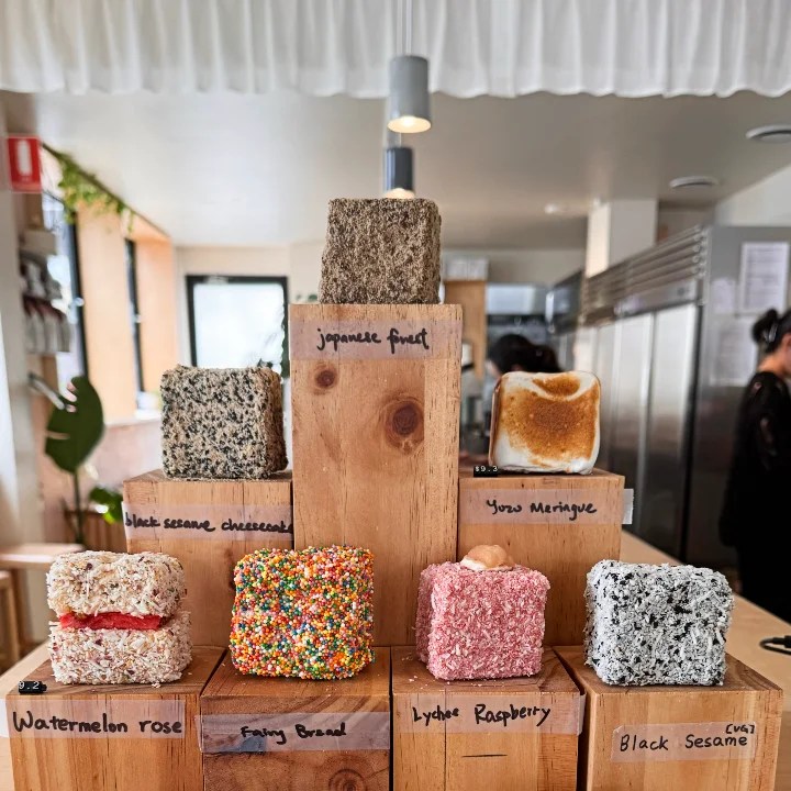 A display of colorful desserts including various flavored lamingtons, arranged in a tiered manner on wooden stands with labels indicating their names, such as 'Watermelon Rose', 'Fairy Bread', and 'Black Sesame'.