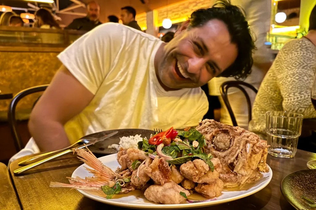 A person smiling while sitting at a table, with a plate of vibrant food that includes a fried whole fish and garnishes, alongside a glass of red wine.