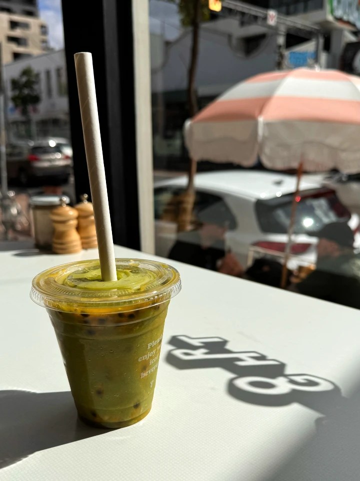 A refreshing green smoothie in a clear cup with a paper straw, sitting on a table with sunlight casting shadows and a striped umbrella visible in the background.
