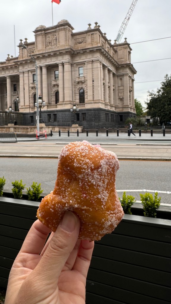 A hand holding a sugar-coated hot jam doughnut in front of a historic building in Melbourne, Australia.