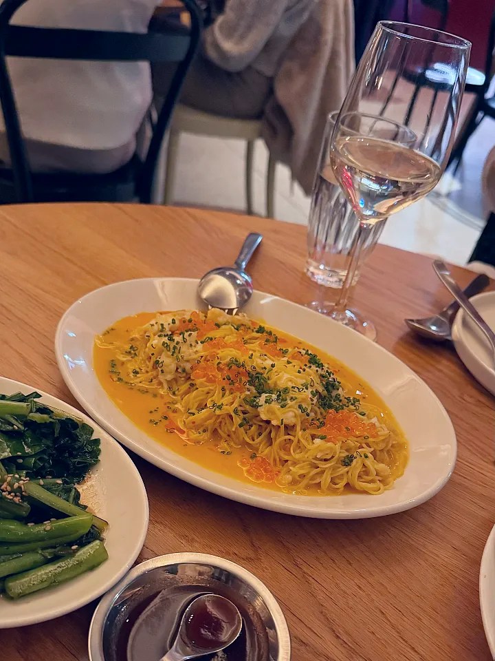 A plate of creamy pasta topped with chives and fish roe, accompanied by a side of sautéed greens and a glass of white wine on a wooden table.