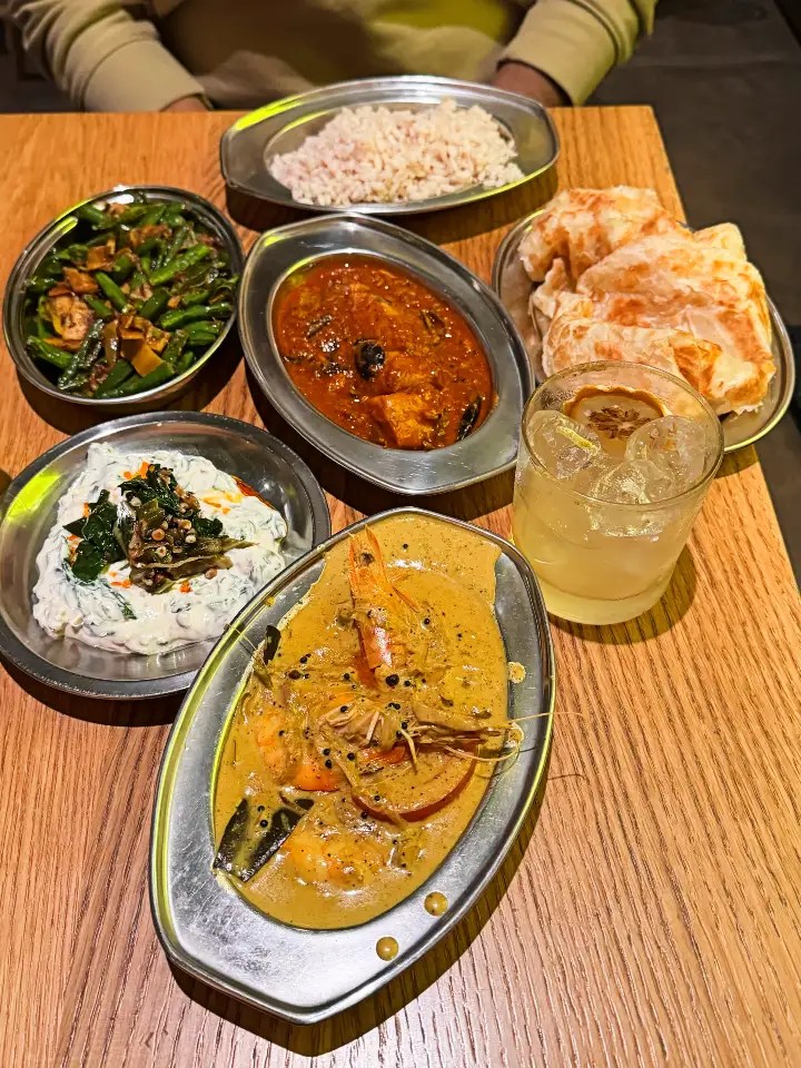 A meal of Indian cuisine featuring a variety of dishes including curry with shrimp, vegetables, rice, roti, and a refreshing drink on a wooden table.