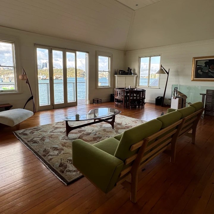 Interior of a boathouse in Sydney featuring large windows with views of the water, a green sofa, and a coffee table on wooden flooring.