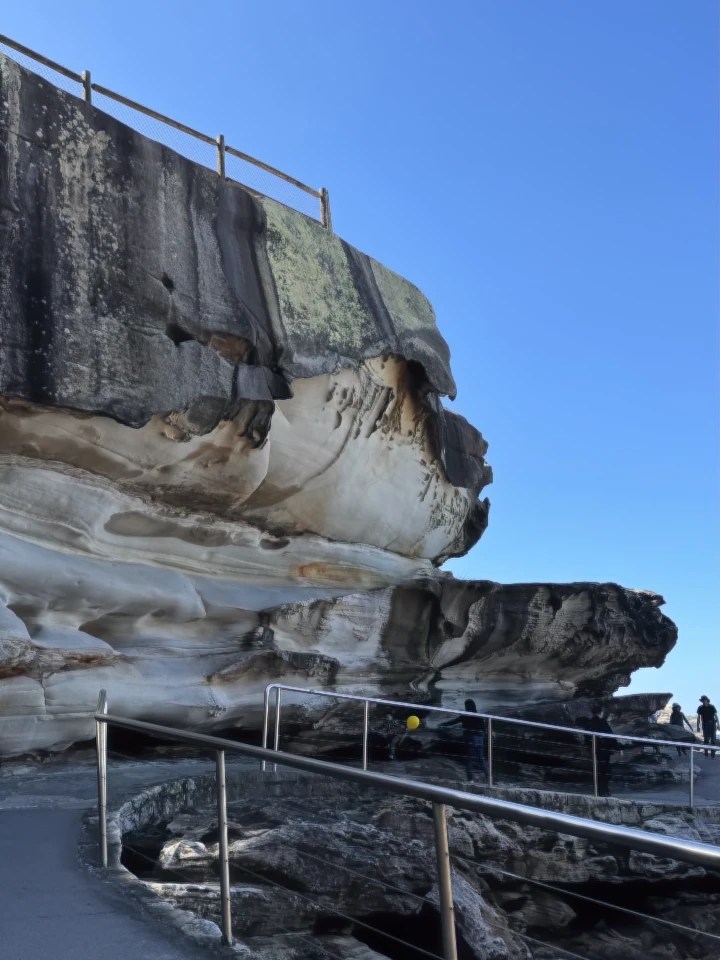 A view of a rocky coastal cliff with a railing along a path, under a clear blue sky.