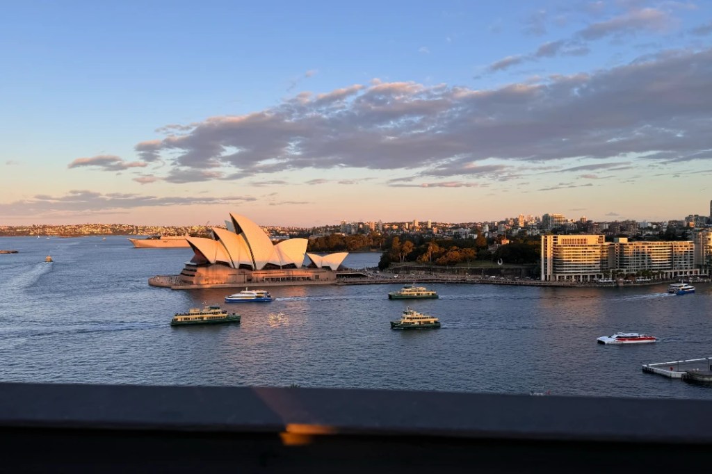 View of the Sydney Opera House from afar during sunset, with ferries passing in the water and a city skyline in the background.