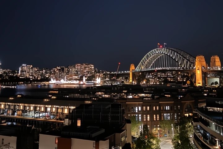 A nighttime view of Sydney Harbour showcasing the illuminated Sydney Harbour Bridge and surrounding city skyline, with waterfront buildings in the foreground.