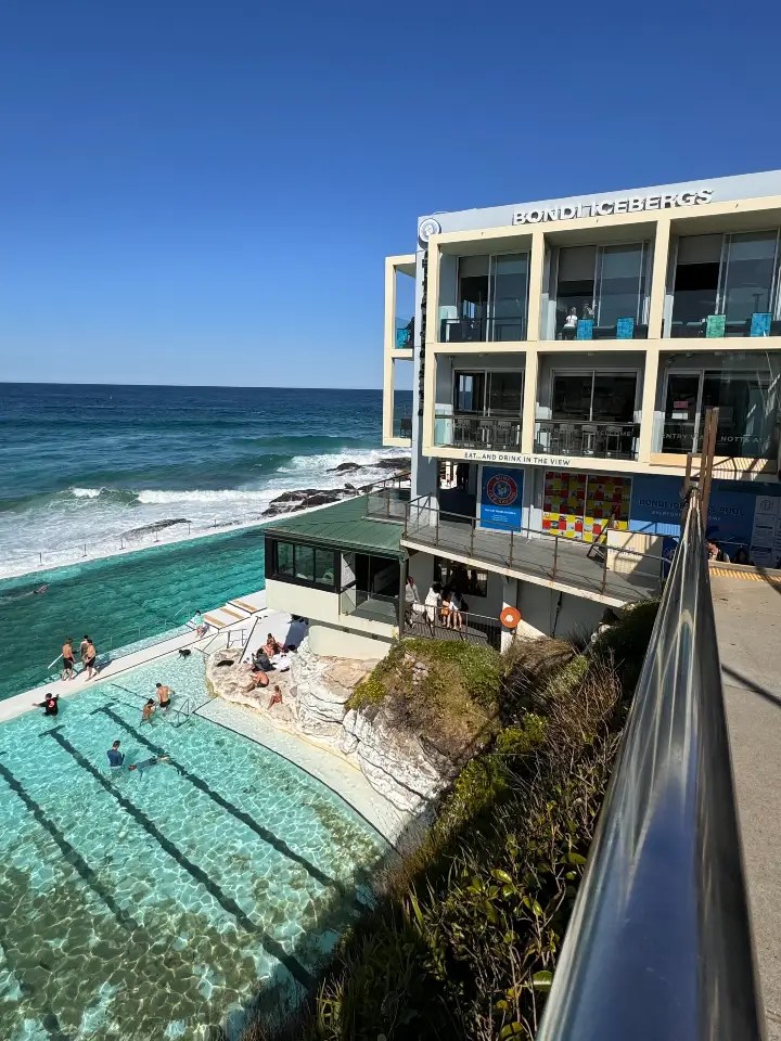 A view of Bondi Icebergs Club, showcasing its ocean pool and nearby rocky coastline, with people enjoying the water on a sunny day.