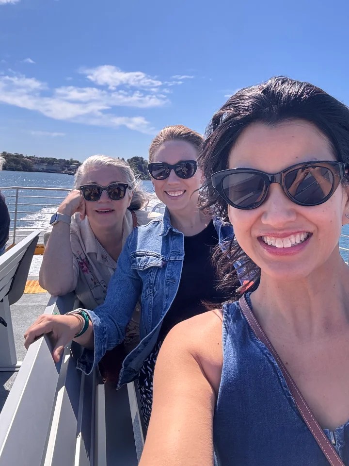 Three women enjoying a ferry ride in Sydney, with a scenic waterfront in the background, under a clear blue sky.
