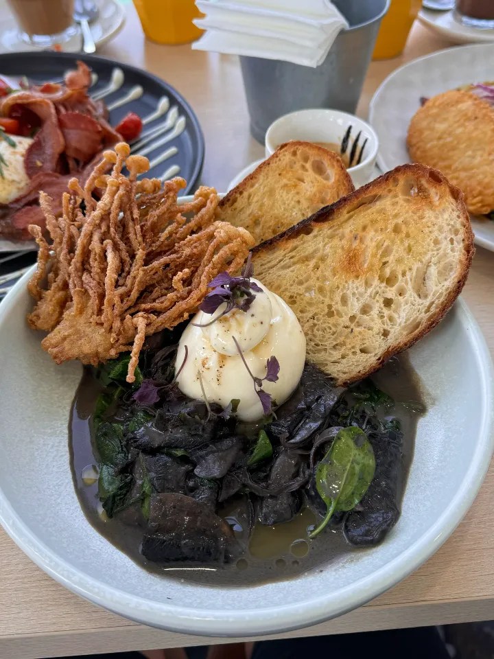 A beautifully plated breakfast dish featuring crispy fried mushrooms, poached egg, sautéed greens and mushrooms and toasted bread on a light-colored plate.