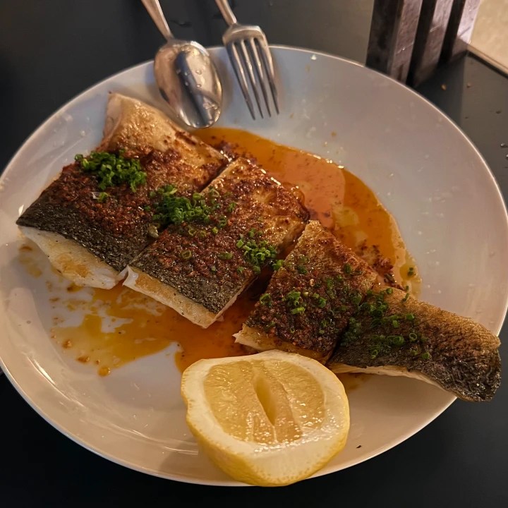 A plate featuring grilled fish with crispy skin, garnished with chopped herbs and served with a lemon wedge.