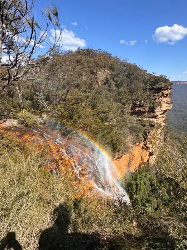 A scenic view of a waterfall cascading down a rocky cliff, with a vibrant rainbow visible in the mist, surrounded by lush greenery and a clear blue sky.