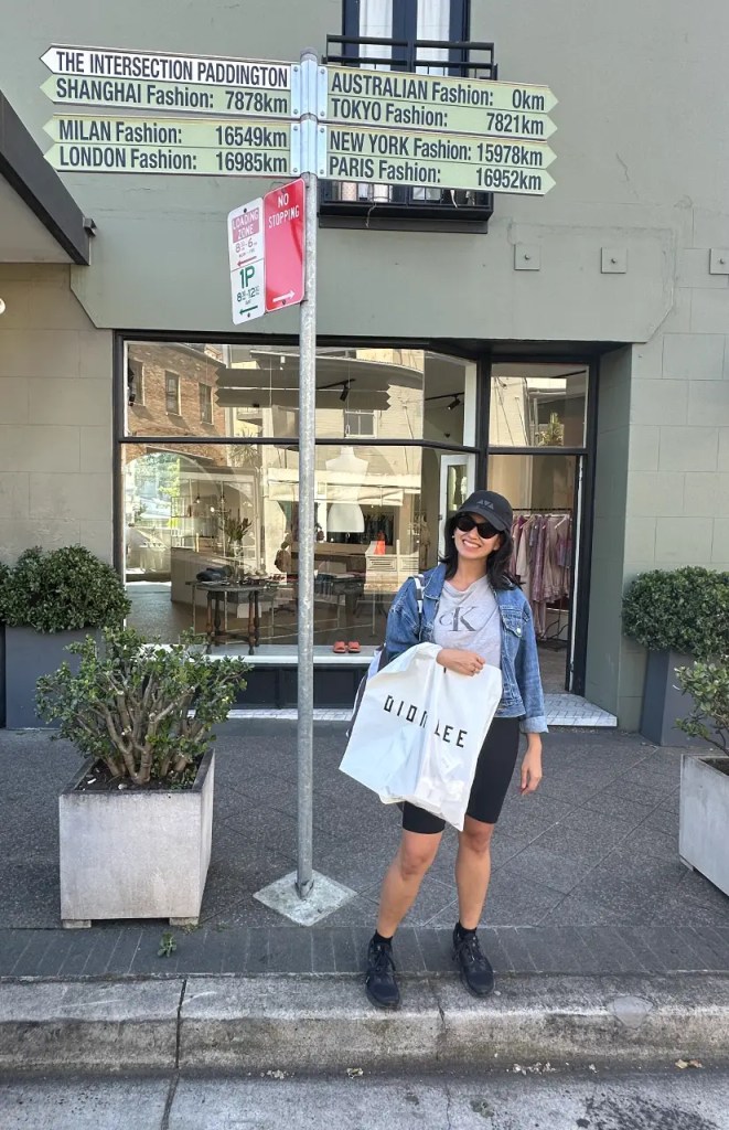 A woman stands at a fashion intersection in Paddington, holding a shopping bag from Dion Lee. The street sign lists various global fashion capitals and distances from Sydney.