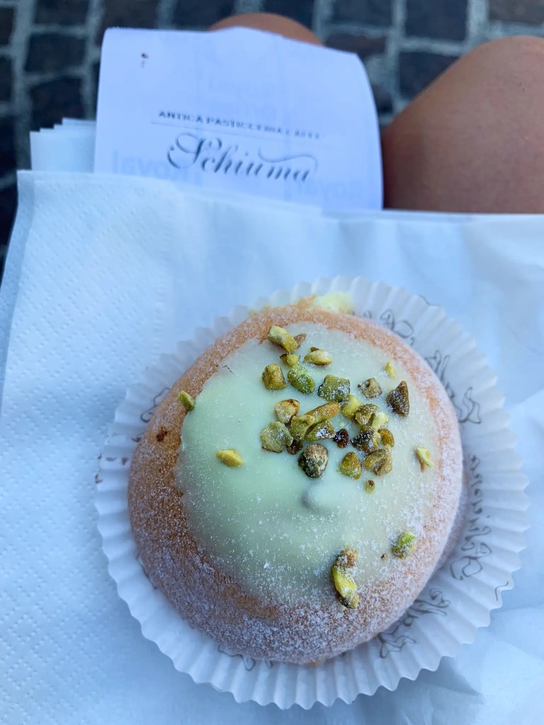 A close-up of a pistachio cream puff topped with chopped pistachios, resting on a white paper napkin, with a receipt from Caffè Schiuma partially visible in the background.