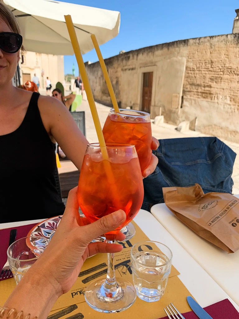 Two glasses of vibrant orange cocktails with straws are clinked together in a celebratory toast at a sunlit outdoor table, with a backdrop of a rustic stone wall.