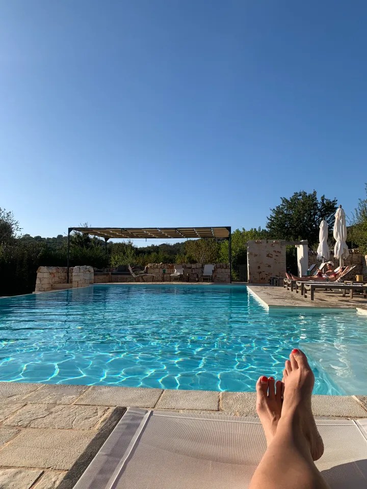 View from a lounge chair by a swimming pool, with bare feet visible in the foreground, clear blue sky above, and sun loungers in the background.