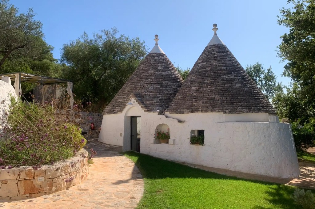 A trullo-style hotel with a conical-roofed stone house, surrounded by greenery with a stone pathway and colorful flowers in the foreground.