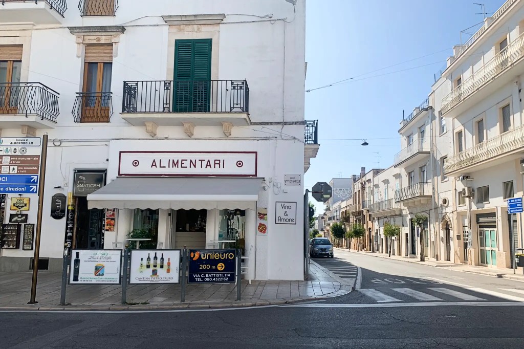 Street view of an alimentari store named 'Vino & Amore' in Alberobello, with a white building featuring balconies and green shutters, alongside a street sign pointing towards Alberobello.
