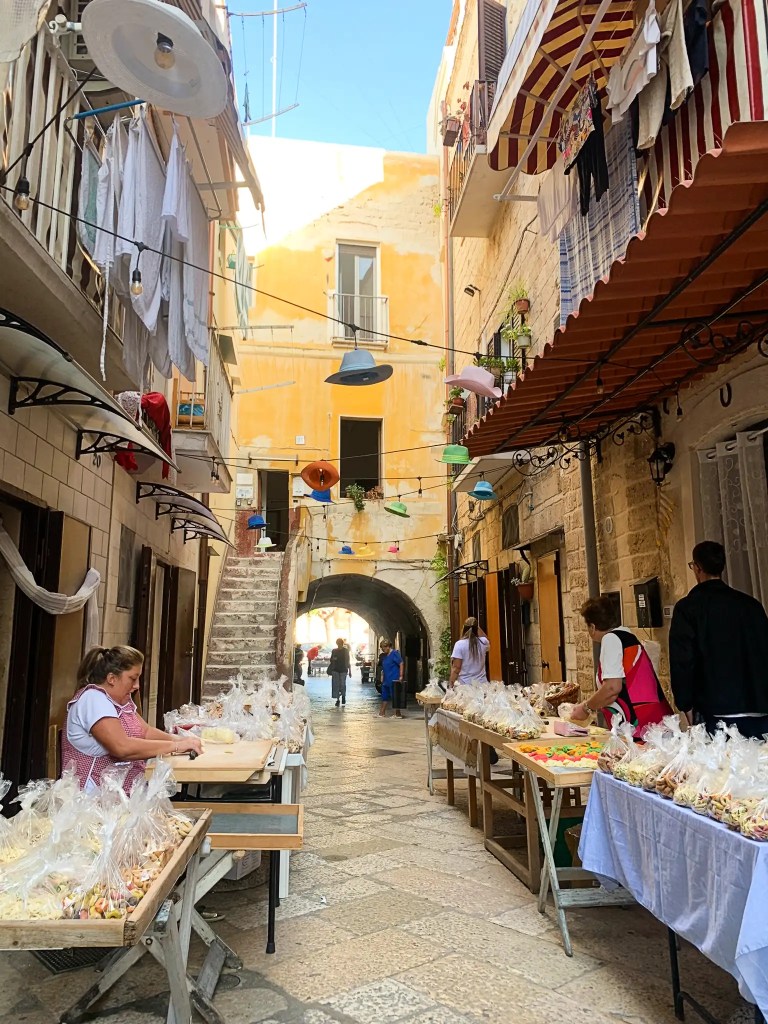 A narrow, bustling street in Bari, Italy with stone buildings, where a woman prepares pasta at a table adorned with colorful treat displays. Strings of lights hang above, and locals stroll by amidst laundry hanging out to dry.