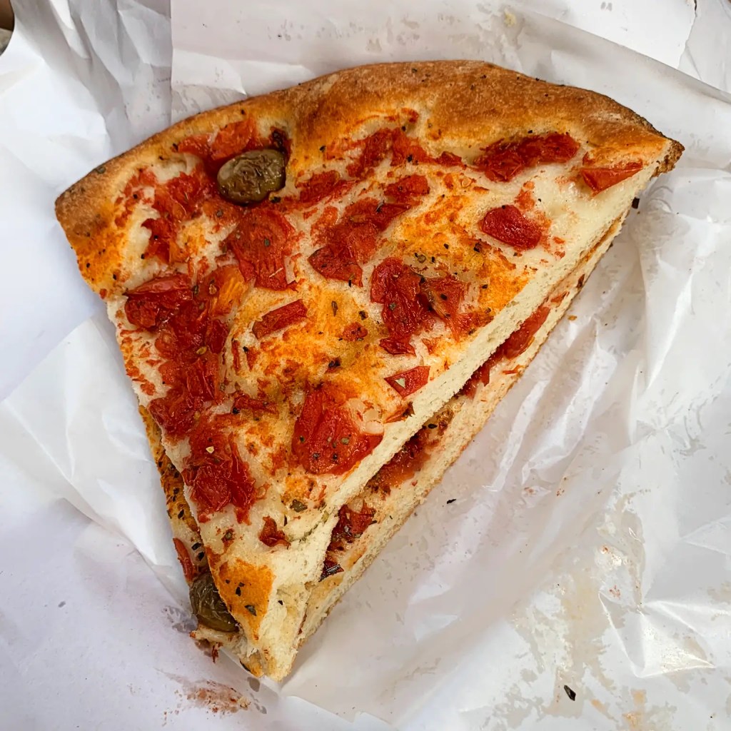 A slice of focaccia Barese topped with tomatoes and olives, wrapped in paper.