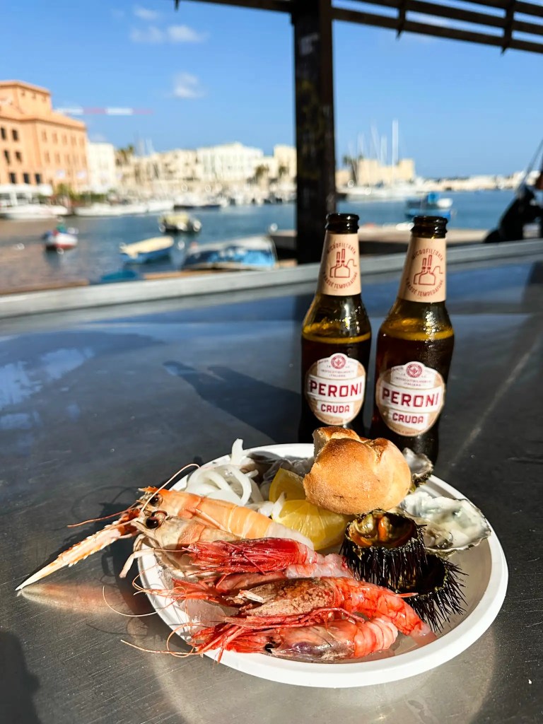 A plate of fresh seafood including shrimp, octopus, and sea urchin, served with a lemon wedge and a small bread roll, alongside two bottles of Peroni beer. In the background, a picturesque harbor with boats and buildings under a clear blue sky.