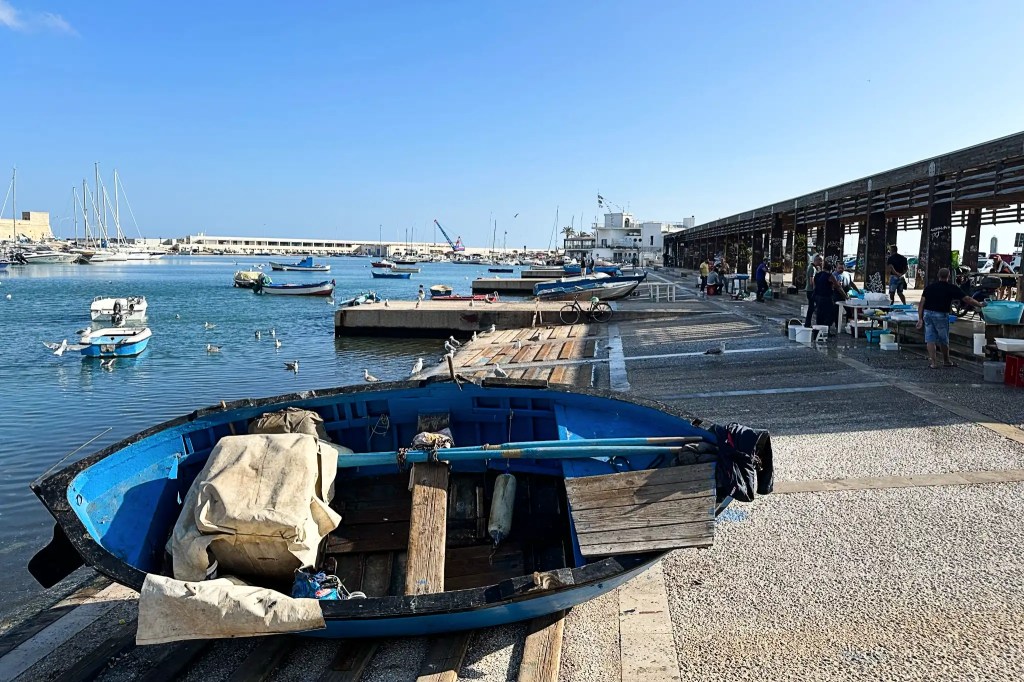 A bright and sunny view of a harbor with small fishing boats docked alongside a stone pier, where people are engaged in activities, under a clear blue sky.