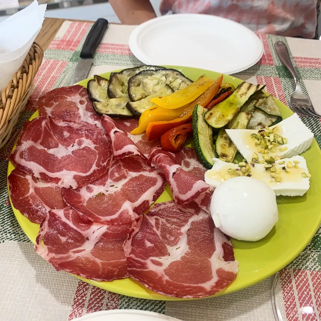 A colorful plate featuring sliced cured meats, including capocollo, complemented by grilled vegetables like eggplant, zucchini, and bell peppers, alongside fresh mozzarella and a creamy burrata topped with pistachios, set on a vibrant yellow dish.