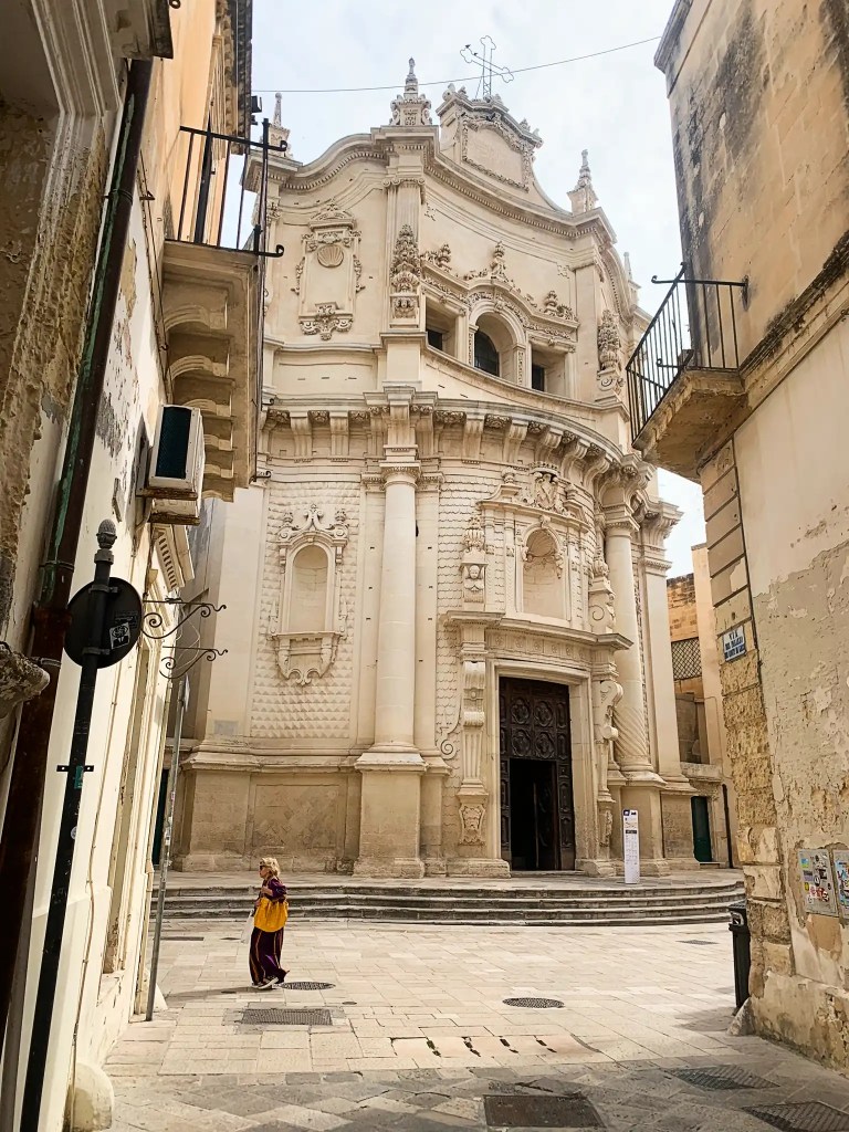 A woman in a long, colorful outfit walks past an ornate baroque church, characterized by intricate details and statues on its façade, located in a narrow street.