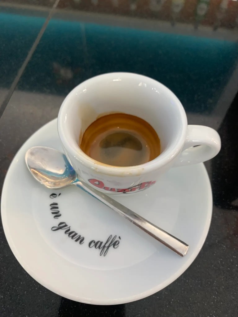 A close-up view of a small white espresso cup on a saucer, with a stainless steel spoon placed beside it. The cup contains a shot of espresso, and the saucer has the text 'è un gran caffè'.