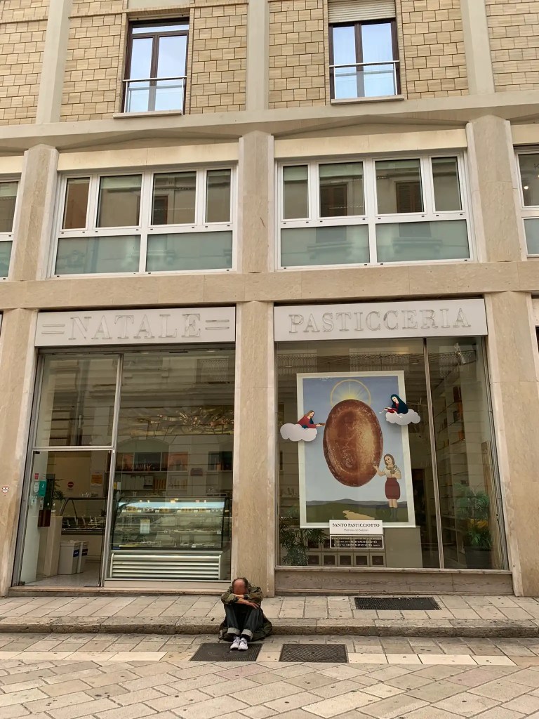 A street view of Natale Pasticceria, a pastry shop in Italy, featuring a large poster of a pastry in the window. A person is sitting on the sidewalk in front of the shop, partially hidden, resting against the building.