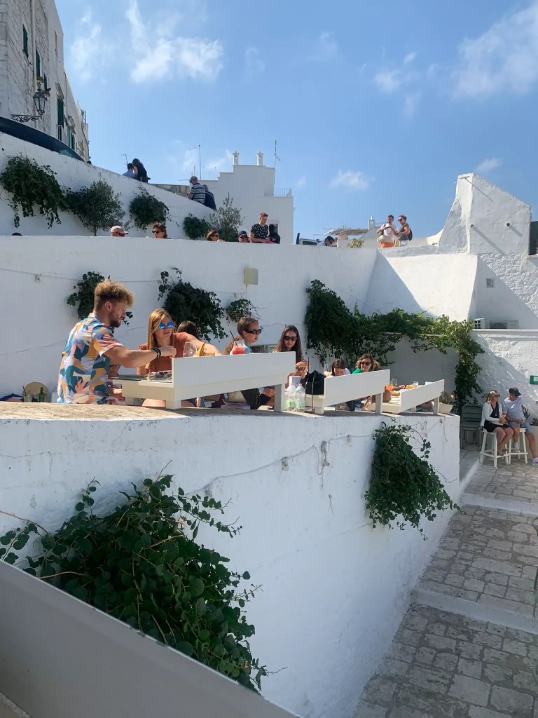 A group of people enjoying food and drinks outside on a white terrace in a sunny location, surrounded by whitewashed buildings and greenery.