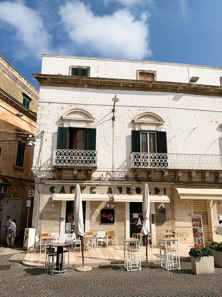 A quaint café in a sunlit Piazza, featuring whitewashed walls, green shutters, and outdoor seating with empty tables and umbrellas.