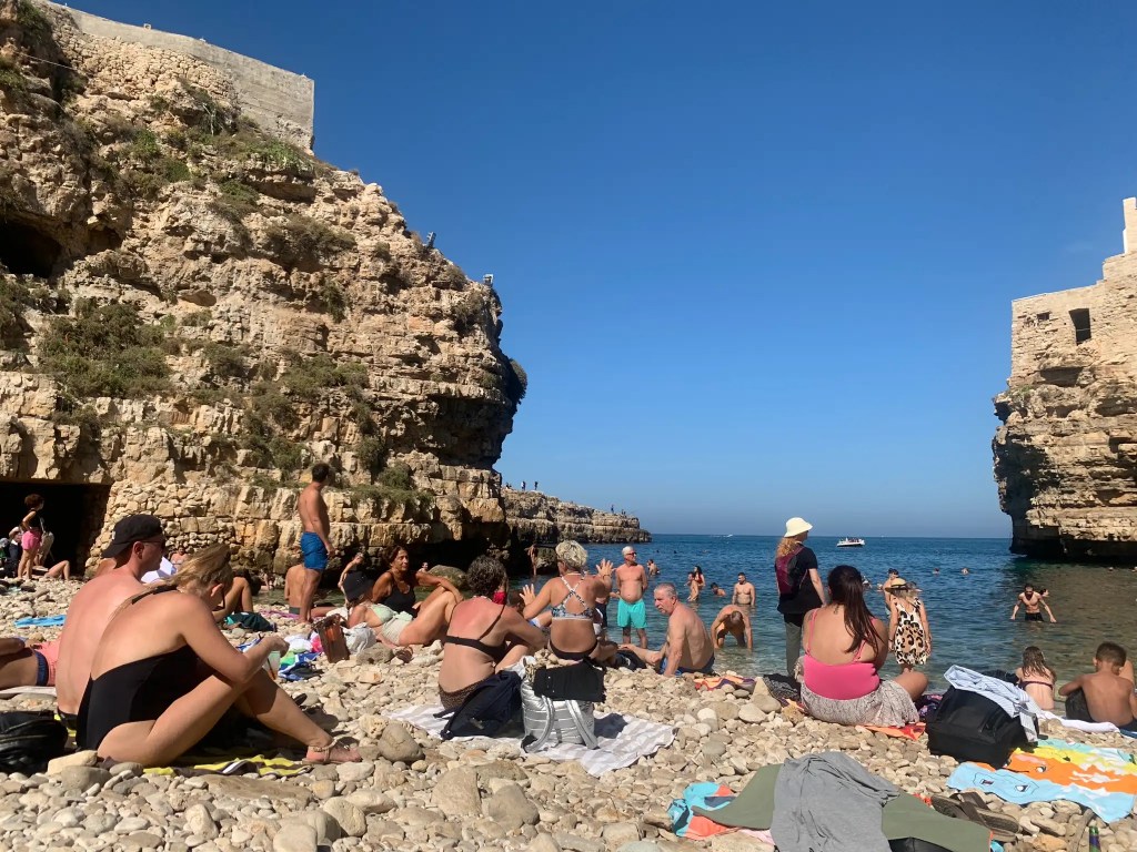 A crowded beach scene in Polignano a Mare, with people sunbathing on pebbles, swimming in the clear blue water, and rocky cliffs in the background.