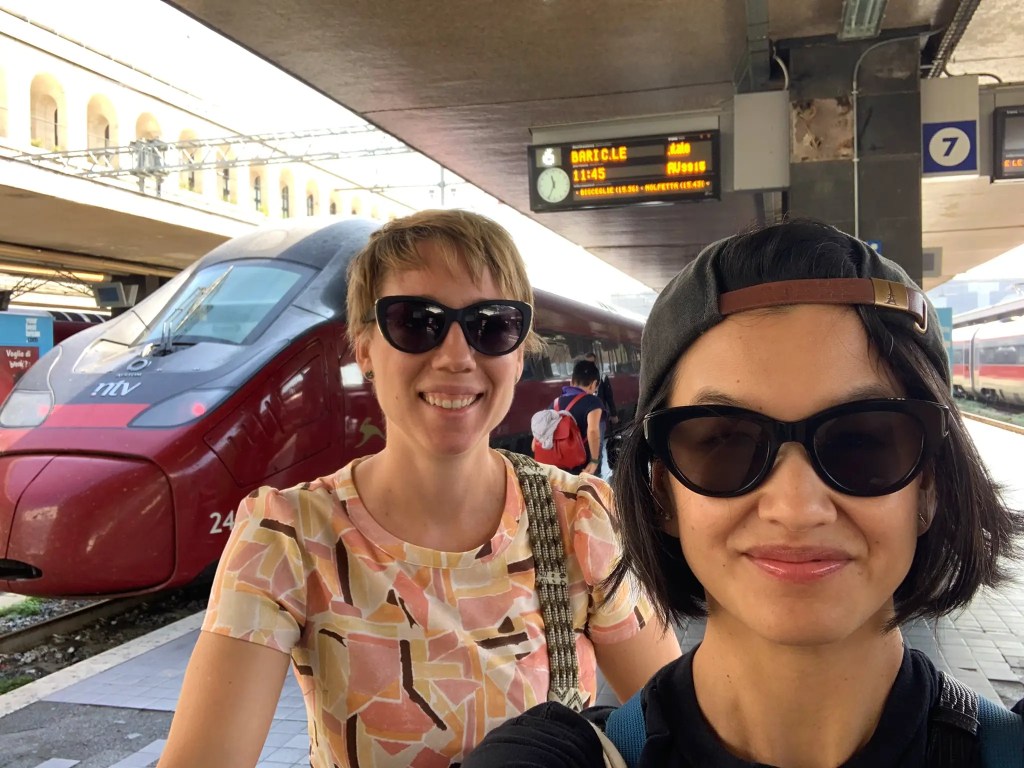 Two women pose for a selfie at a train station in Italy, with a red high-speed train visible in the background and a departure board showing schedules.