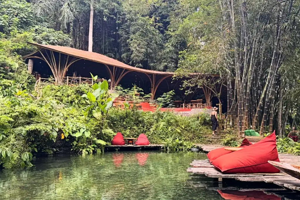 A serene eco-lodge nestled among lush greenery in Ubud, Bali, featuring a wooden restaurant with a sloping bamboo roof and red outdoor bean bags, surrounded by vibrant plants and a natural swimming pool.
