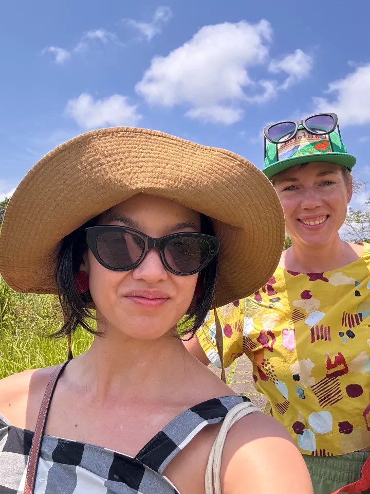 Selfie of two women smiling outdoors, wearing sunglasses and hats, with a bright sky in the background.