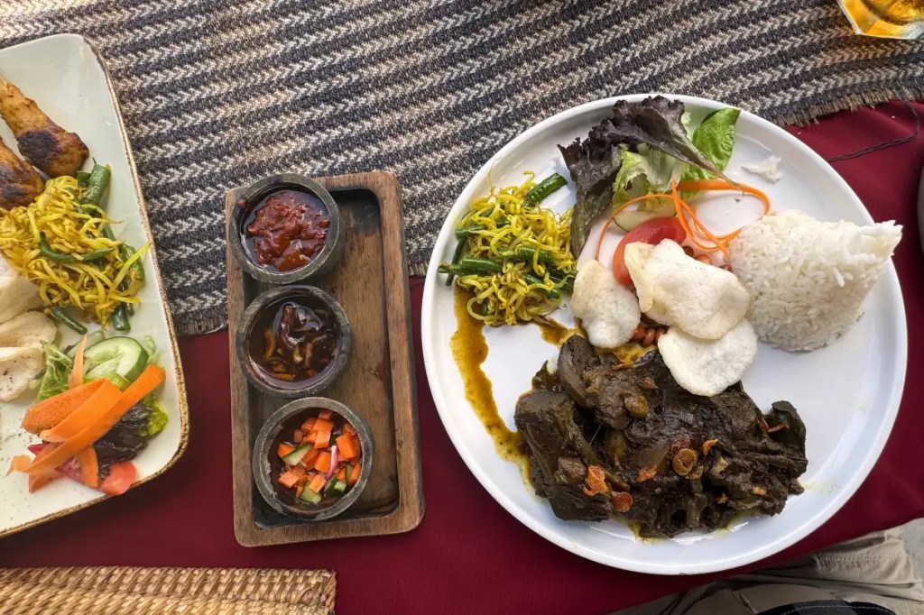 A plate of traditional Indonesian food featuring jackfruit rendang, rice, and a variety of side dishes including salad, vegetables, and crackers, accompanied by small bowls of sauce.