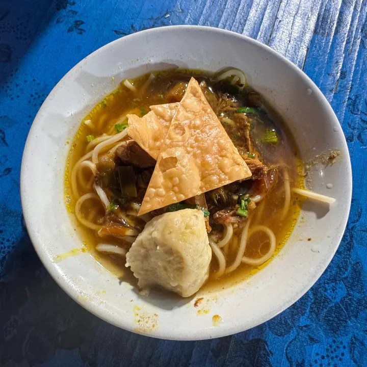 A bowl of Indonesian noodle soup topped with crispy crackers, served on a blue table.