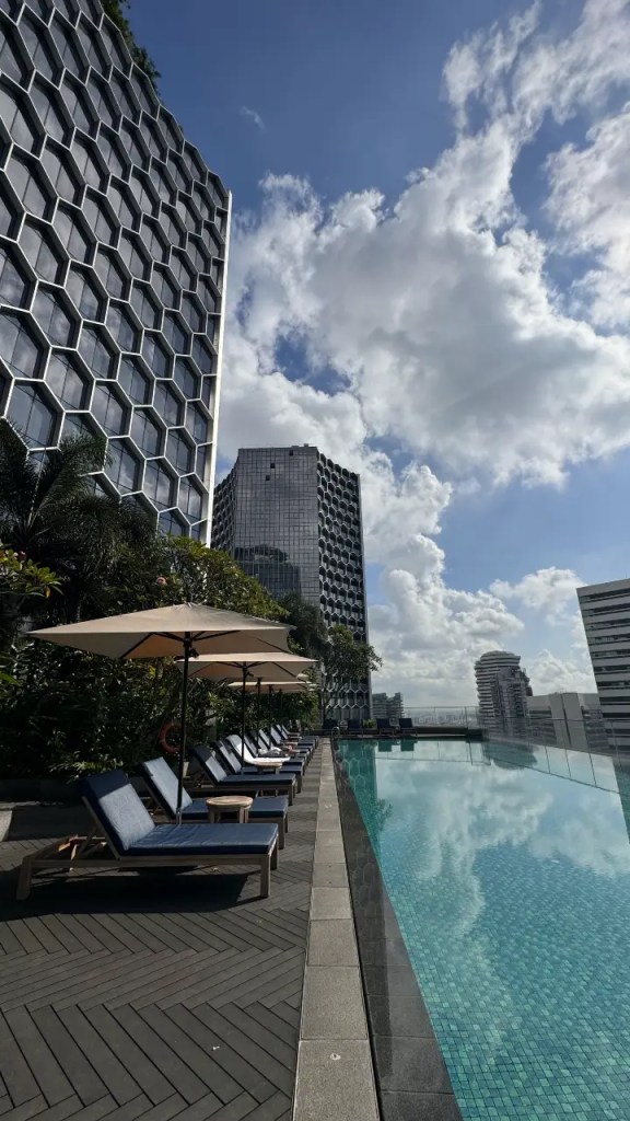 An outdoor rooftop infinity pool with lounge chairs, surrounded by modern buildings under a blue sky with fluffy clouds.