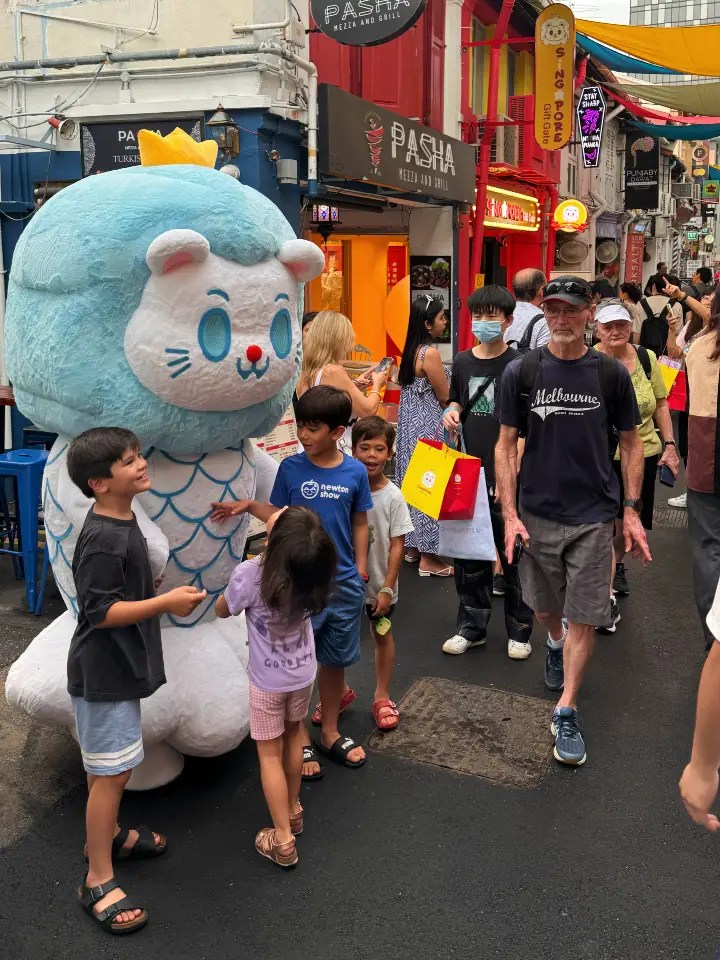 A colorful street scene in Singapore featuring a Merlion character surrounded by children, with shops and people in the background.