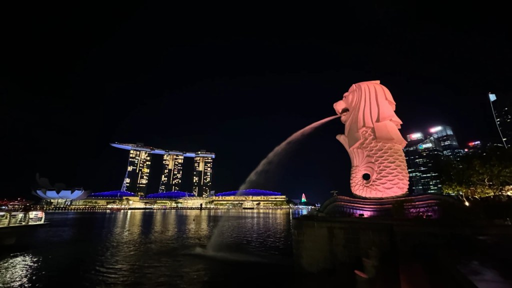 The Merlion statue spouting water at Marina Bay, illuminated with pink lights against the night sky, with the Marina Bay Sands hotel in the background.