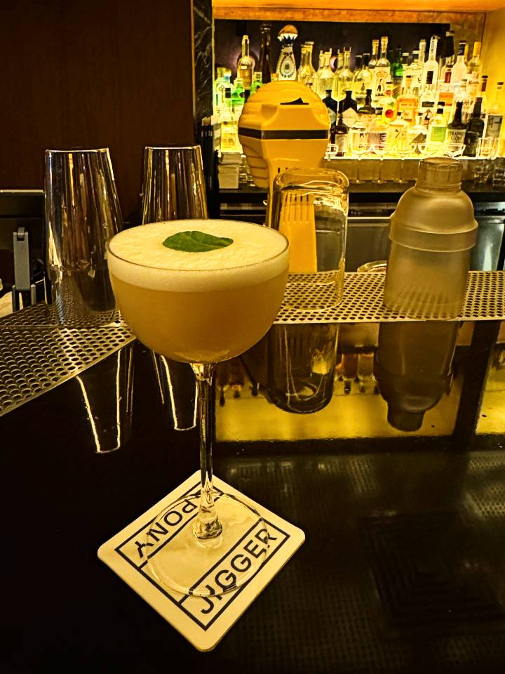 A cocktail with a foamy top is served in a stylish glass at a bar, with a blurred background of various liquor bottles.
