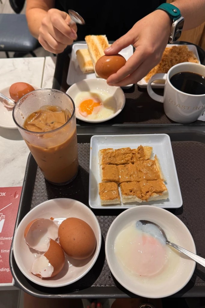 A tray featuring breakfast items: two soft-boiled eggs, toast with peanut butter, another piece of toast, a bowl of soft-boiled eggs, a glass of iced coffee, and a cup of black coffee.
