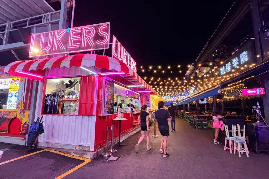 Night view of a colorful food stall named 'LUKERS' with bright neon lights, surrounded by patrons enjoying the outdoor dining space under string lights.