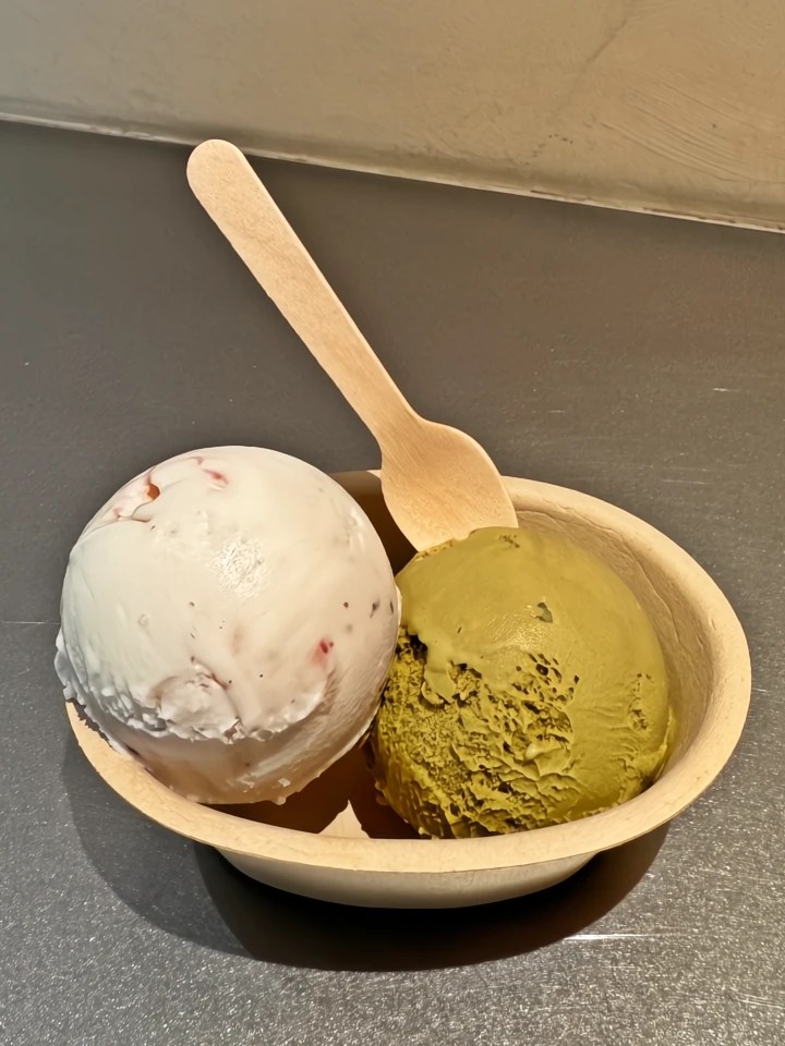A wooden bowl containing two scoops of gelato: one white with strawberry swirls and one green, matcha flavor, accompanied by a small wooden spoon.