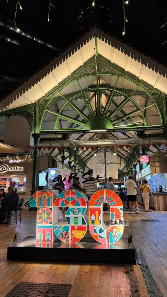 Illuminated sign celebrating 130 years of Lau Pa Sat, a historic food market, with decorative motifs and people enjoying the atmosphere in the background.