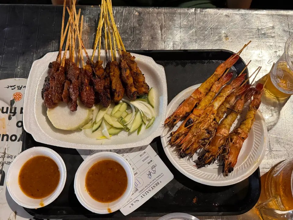 A platter featuring skewers of grilled satay with a serving of grilled prawns, accompanied by slices of cucumber and rice cakes, and two small bowls of dipping sauce.