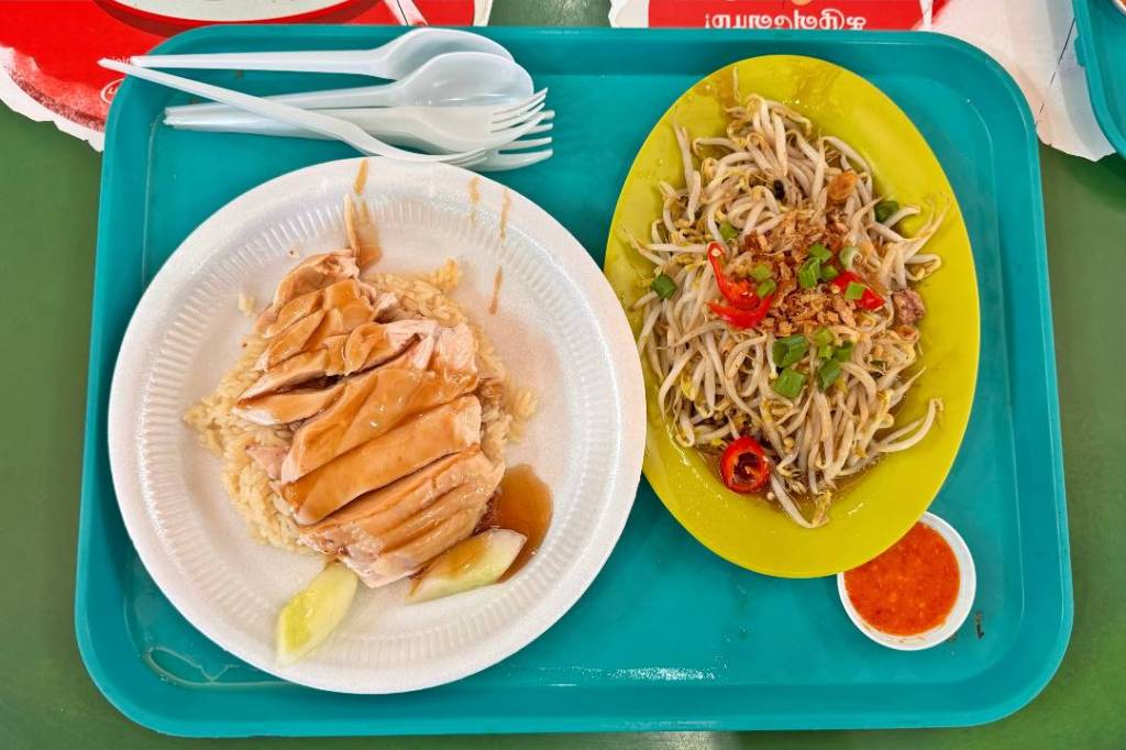 A serving of Hainanese chicken rice on a white plate, with slices of chicken drizzled in sauce, accompanied by slices of cucumber and a small bowl of chili sauce, next to a plate of bean sprouts with green onions and red chili peppers, all placed on a teal tray.